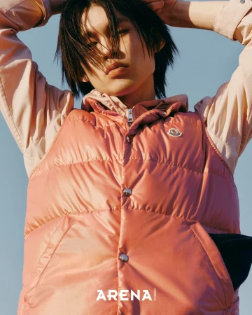 Model wearing a pink puffer vest and light pink hoodie with hands behind head against a clear sky background