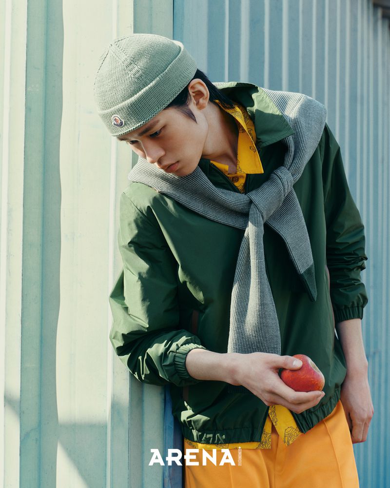 Model wearing a green beanie, dark green jacket, yellow shirt, and holding an apple against a corrugated metal wall.