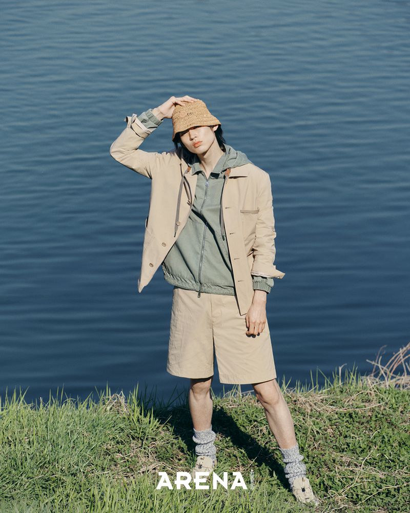 Model wearing tan jacket and shorts holding bucket hat by water for Arena Homme Plus Korea