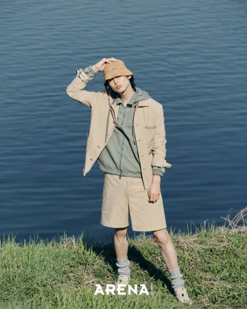 Model wearing tan jacket and shorts holding bucket hat by water for Arena Homme Plus Korea