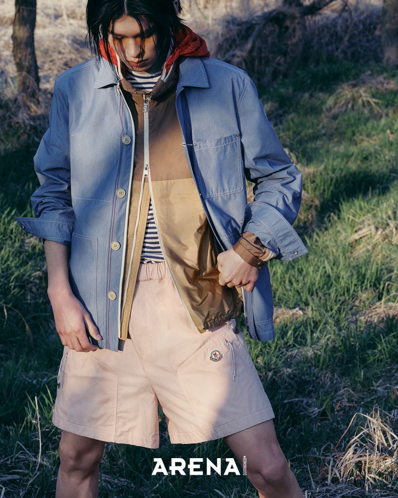 Model in blue jacket and light pink shorts standing outdoors in a grassy field with trees under natural light