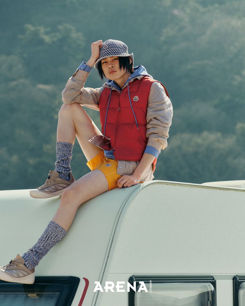 Male model sitting on a vehicle roof wearing a red vest, yellow shorts, patterned bucket hat, and high socks outdoors