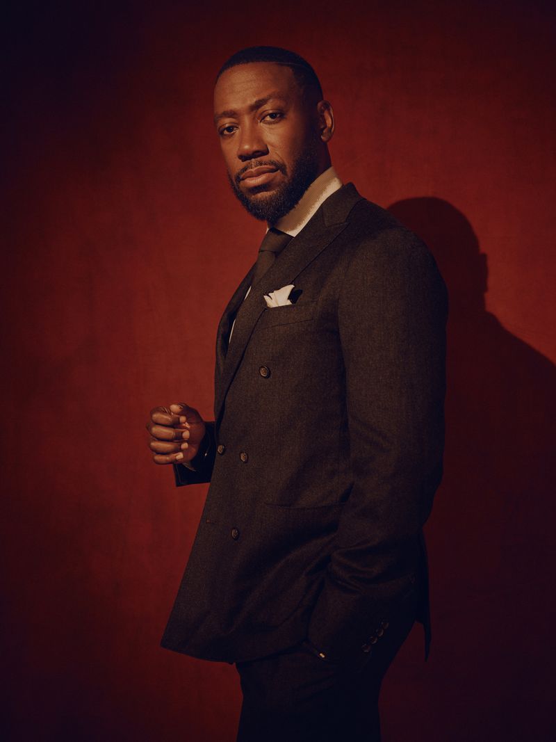 Portrait of Lamorne Morris wearing a dark double-breasted suit against a textured red background
