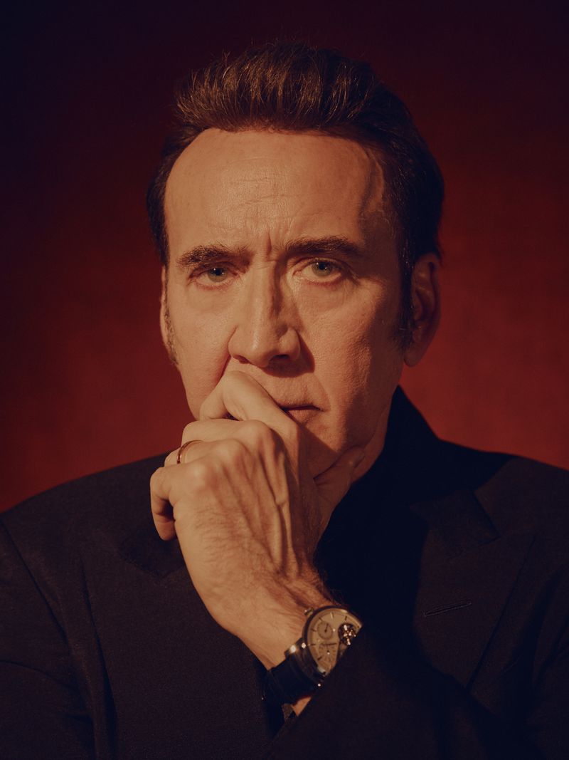 Portrait of Nicolas Cage with hand to his chin, wearing a dark suit and wristwatch against a dark red background.