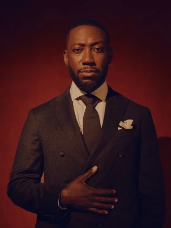 Portrait of Lamorne Morris wearing a dark double-breasted suit and tie against a deep red background.