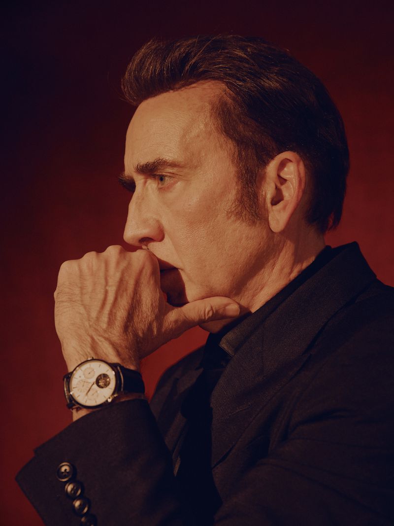 Profile portrait of Nicolas Cage with hand on chin wearing a dark jacket and watch against a warm red background.