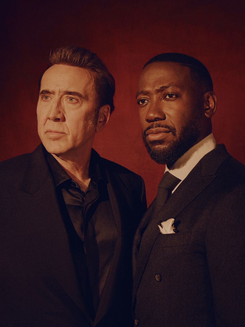Portrait of Nicolas Cage and Lamorne Morris wearing dark suits against a textured red background photographed by David Urbanke for LA Times.