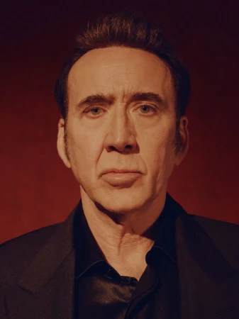 Portrait of actor Nicolas Cage with a serious expression, wearing a black suit jacket against a dark red background in a studio setting.