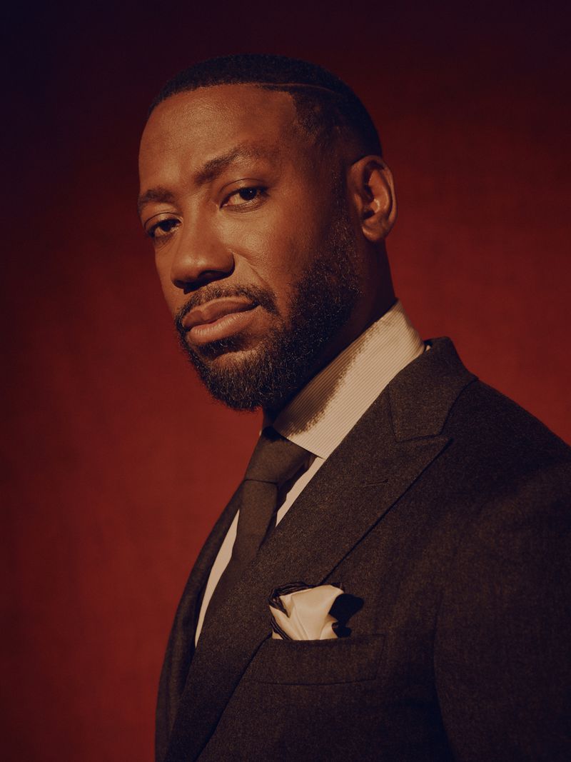 Portrait of Lamorne Morris wearing a dark suit and tie against a warm red textured background