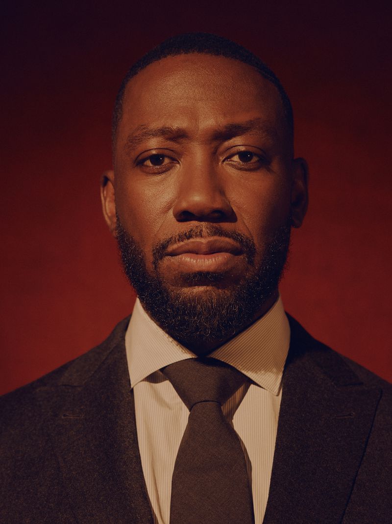 Portrait of Lamorne Morris wearing a dark suit and tie against a warm red background for the LA Times by David Urbanke.