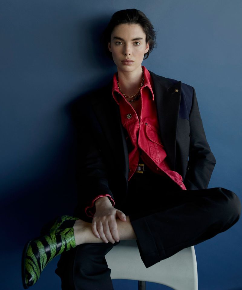 Model wearing a black blazer over a red collared shirt and green patterned boots, seated against a dark blue wall.