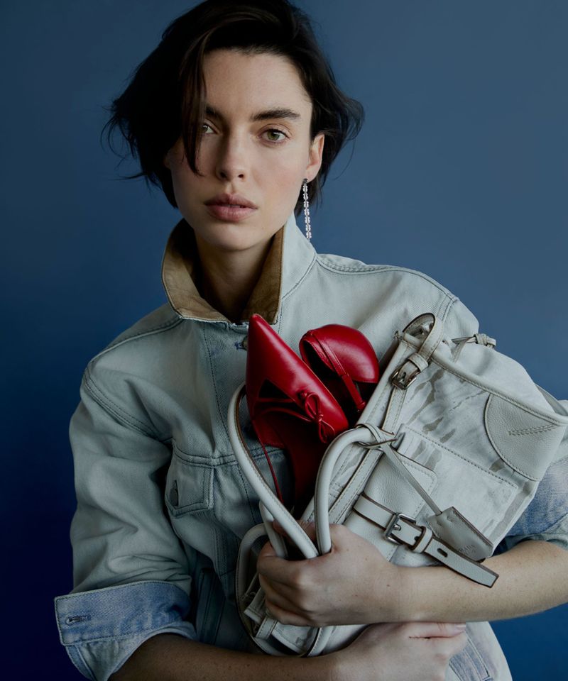 Portrait of a woman wearing a denim jacket holding a cream-colored handbag and a pair of red high-heeled shoes indoors.