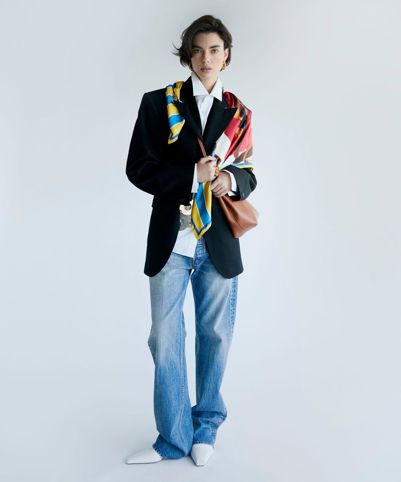 Model wearing a black blazer, white shirt, and blue jeans, styled with a colorful patterned scarf and a brown handbag against a white backdrop.