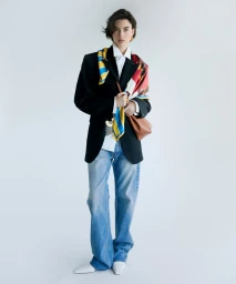 Model wearing a black blazer, white shirt, and blue jeans, styled with a colorful patterned scarf and a brown handbag against a white backdrop.