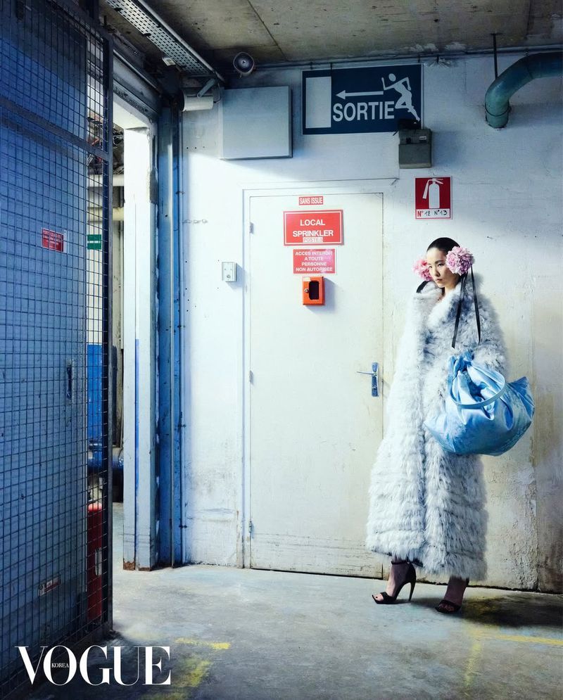 Model wearing a fluffy white coat and blue bag standing in an industrial utility corridor with a 'SORTIE' sign