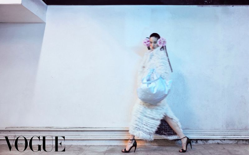 Model wearing a white furry coat and holding a large blue bag against a plain wall for Vogue Korea.