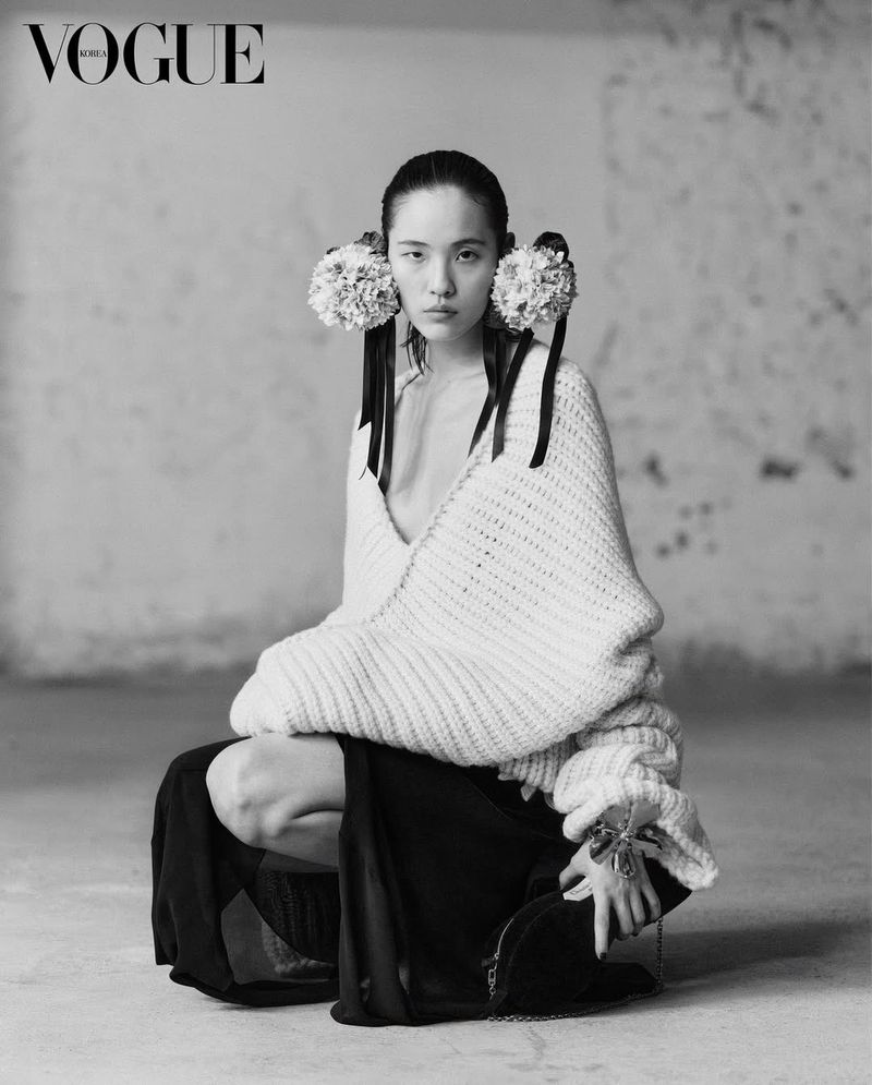 Black-and-white portrait of an Asian model in an off-white knit sweater, large floral hair accessories, and a long black skirt against a distressed wall.