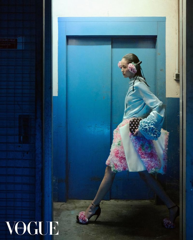 Model in blue silk blouse and floral skirt walking past bright blue elevator doors for Vogue Korea editorial
