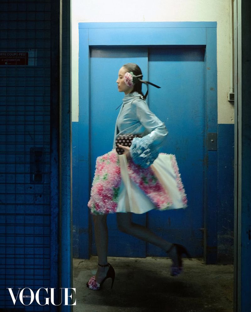 Model in a light blue ruffled top and floral skirt running past a blue elevator door for Vogue Korea.