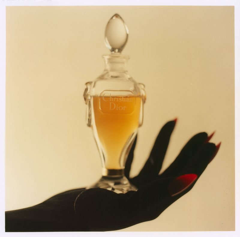 An ornate glass perfume bottle resting on an open gloved hand with red fingernails against a warm background.