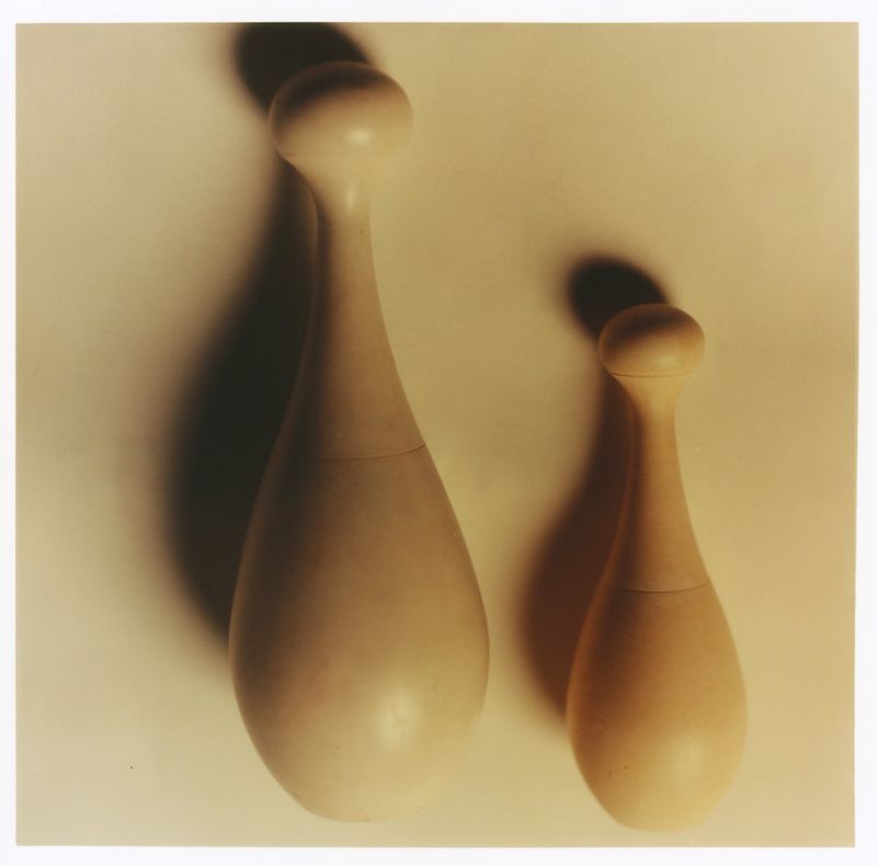 Two smooth wooden bottles or sculptural objects standing upright casting distinct shadows against a light neutral background.