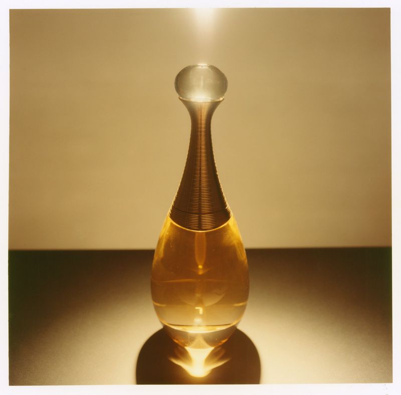 Close-up product shot of a teardrop-shaped perfume bottle with a golden liquid and metallic neck against a dark surface.