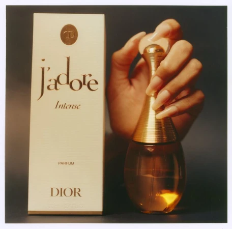Perfume still life with a hand holding a golden bottle of Dior J'adore Intense, next to a box, against a dark background, studio shot.