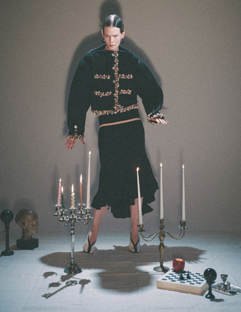 A stylish fashion shot features a woman in a dark jacket and skirt, surrounded by candles and various props on a light neutral background.