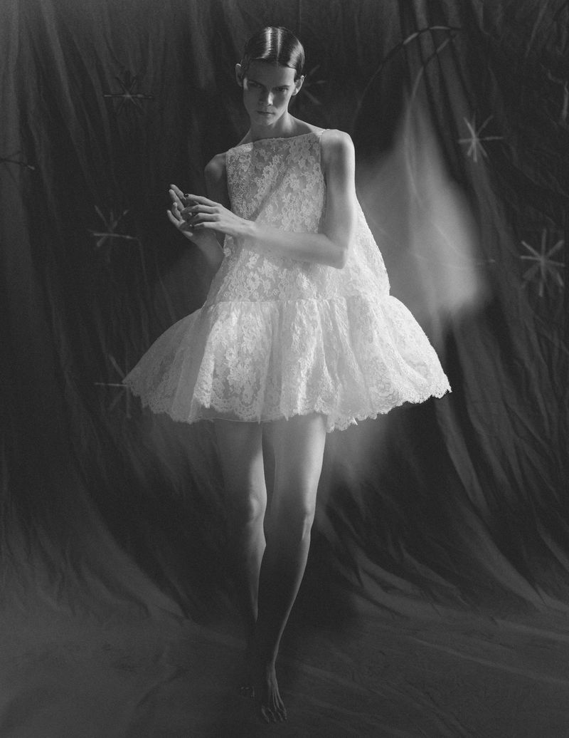 Model Irina Kravchenko in a white lace minidress against a dark textured backdrop for L'Officiel Italia