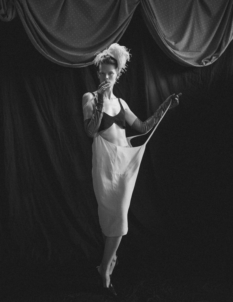 Model Irina Kravchenko in black bra and draped white dress holding strap against dark draped backdrop for L'Officiel Italia