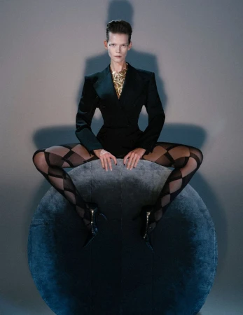 Model Irina Kravchenko wearing a structured black blazer and patterned tights perched on a dark orb.