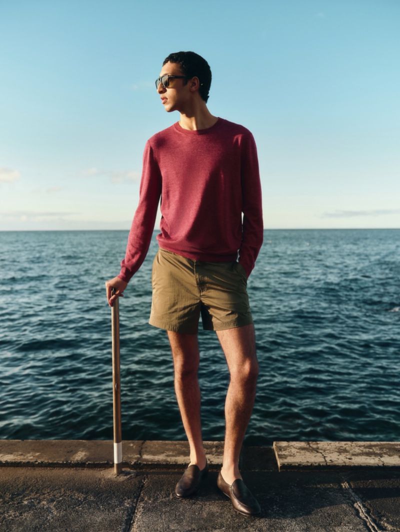 Man wearing a red sweater and shorts standing on a stone pier by the sea under a clear sky during the Mr Porter Off Duty campaign