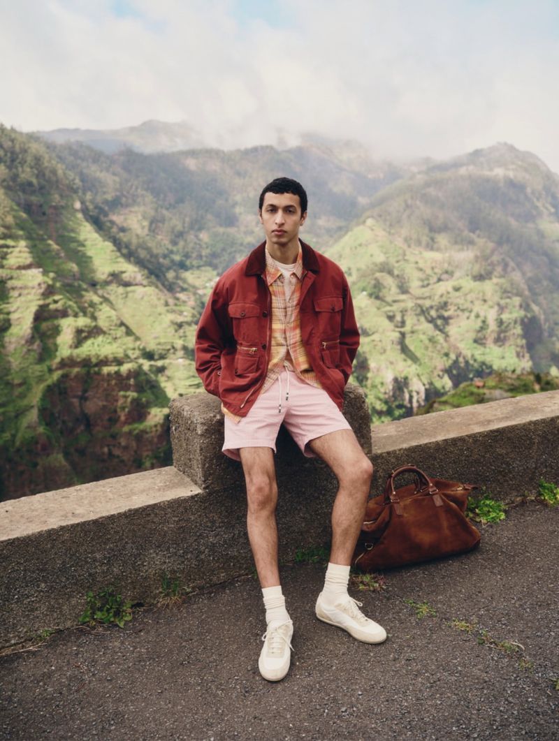 Model wearing a red jacket and pink shorts sitting on a stone wall against a mountain background for Mr Porter.