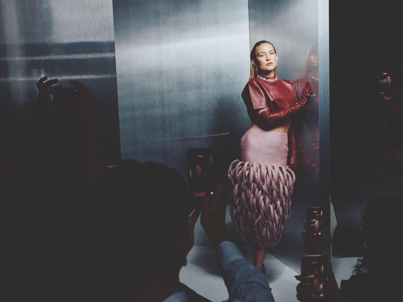 Kate Hudson posing in a red top and textured pink skirt against a metallic background surrounded by people recording with phones.