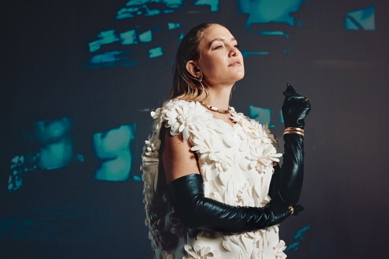Kate Hudson wearing white floral dress and black leather gloves against blue projected background