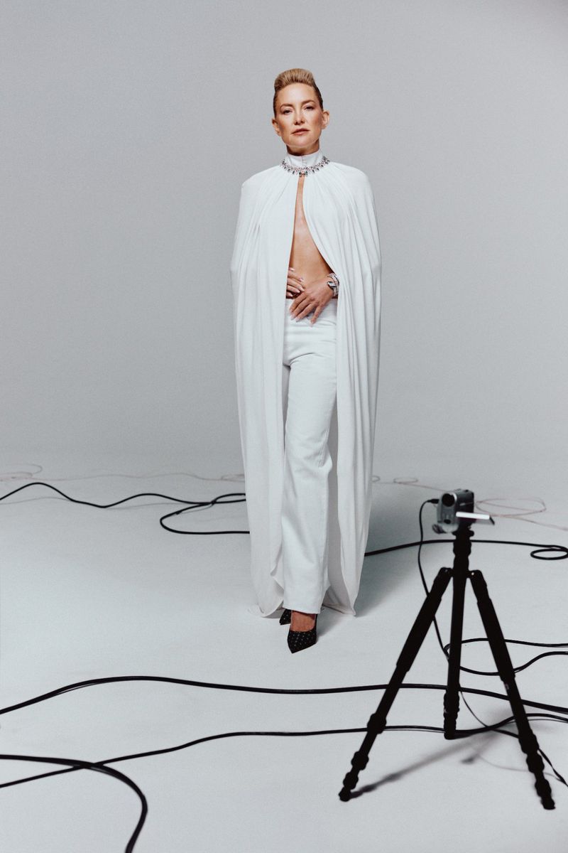 A full-length shot of a woman in an all-white outfit with jewelry and a watch, posing in a photo studio with lighting equipment.