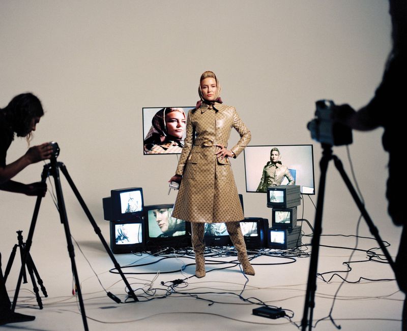 Kate Hudson posing in a patterned trench coat and matching headscarf amid television monitors in a white studio.