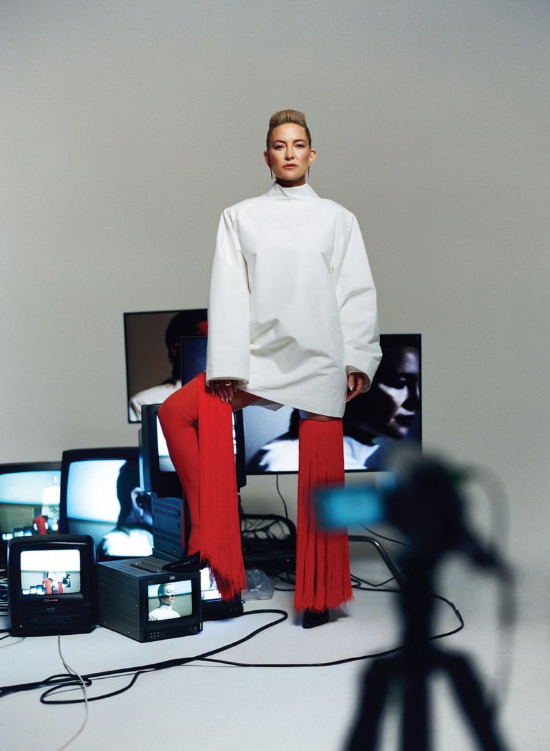 Kate Hudson in a white top and red fringed trousers posing among stacked vintage televisions on a grey seamless background