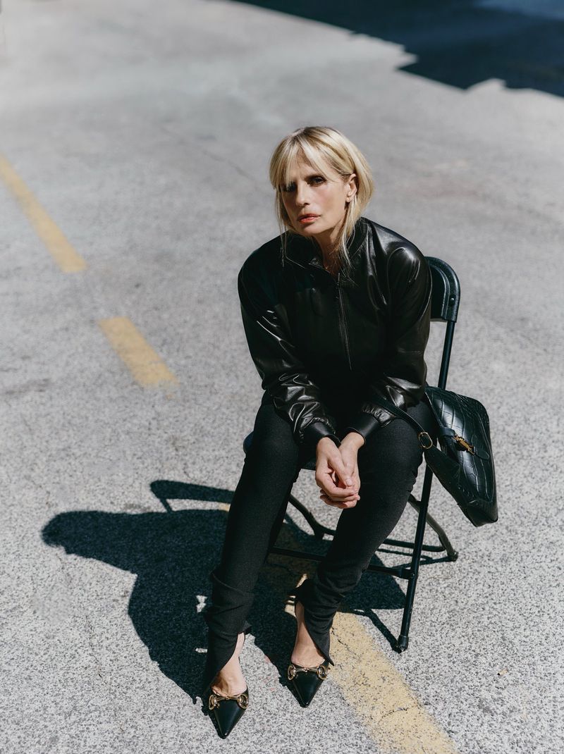 Blonde woman in leather jacket sits on chair on city street in medium shot; black purse, black shoes and clothing, urban environment.