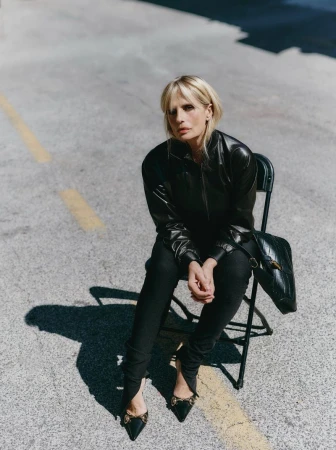 Blonde woman in leather jacket sits on chair on city street in medium shot; black purse, black shoes and clothing, urban environment.