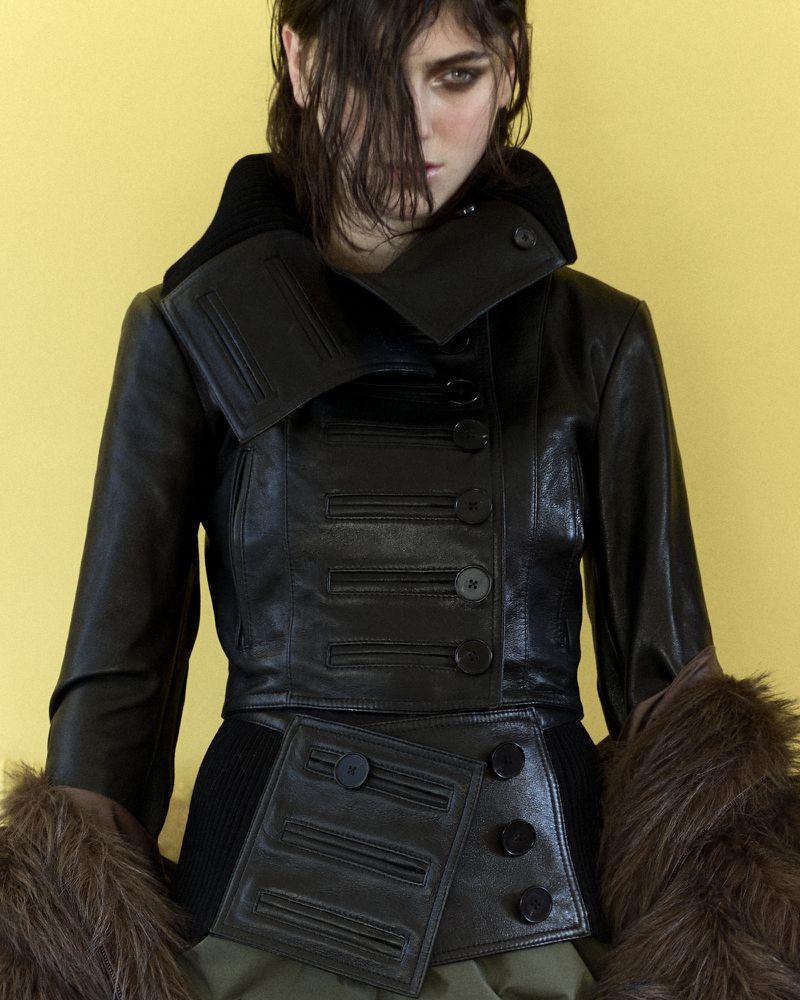 A woman poses indoors wearing a black leather jacket with asymmetrical button design, and fur sleeves against a yellow background.