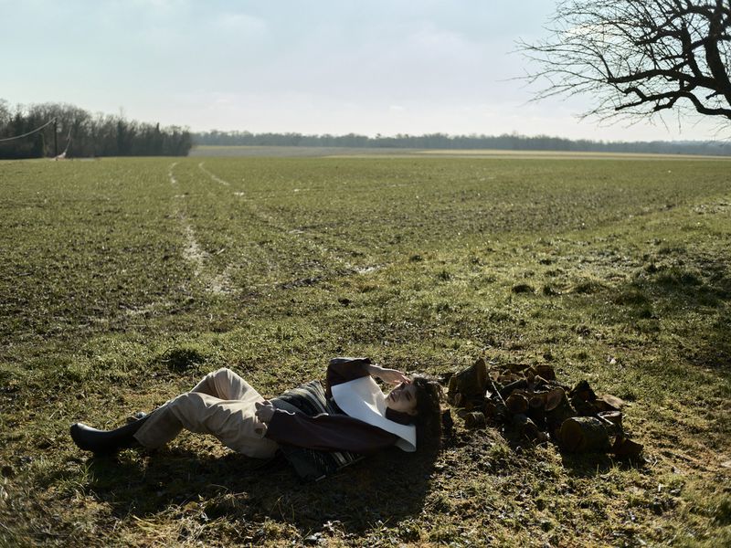 Adult female rests in a field near tree branches, wearing a light top and trousers, under a bright sky on a sunny day in a rural landscape.