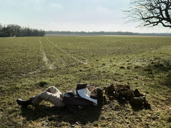 Adult female rests in a field near tree branches, wearing a light top and trousers, under a bright sky on a sunny day in a rural landscape.