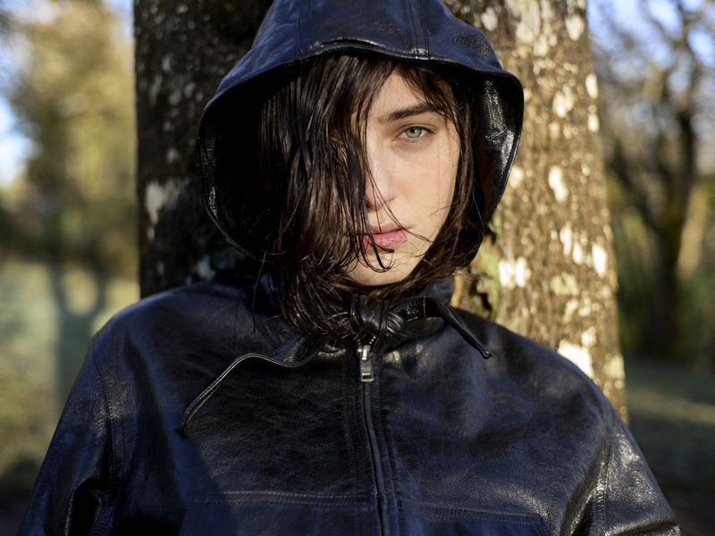 Close-up of model Margaux Martin wearing a black hooded leather jacket leaning against a tree trunk in dappled sunlight.
