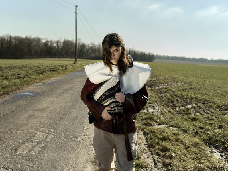 Woman in a brown jacket and white collar stands on a rural road flanked by fields under a bright sky; trees in the distance.
