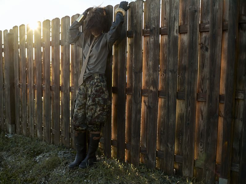 A person leans against a wooden fence in sunlight, wearing a grey shirt, camouflage pants, black boots, and gold bracelets.