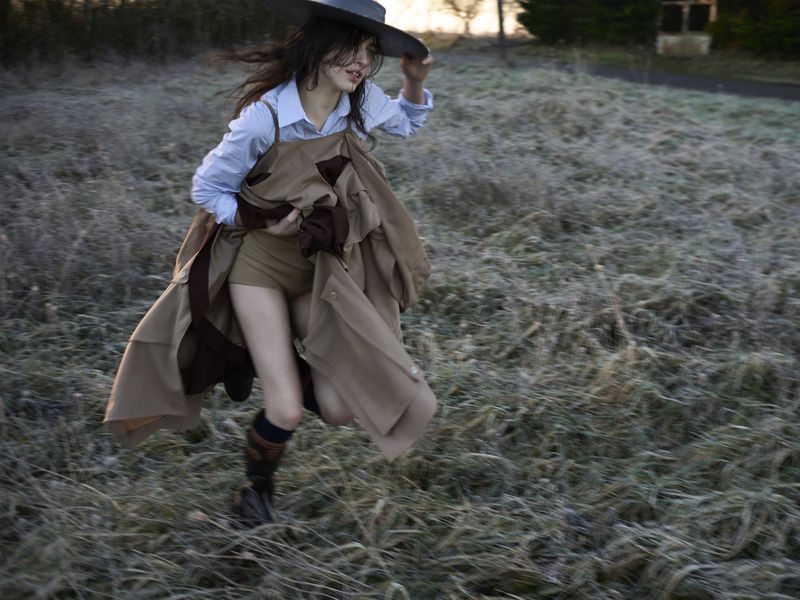 A woman runs through a grassy field wearing a light blue button-down shirt, tan shorts, a brown trench coat, and a wide-brimmed hat.