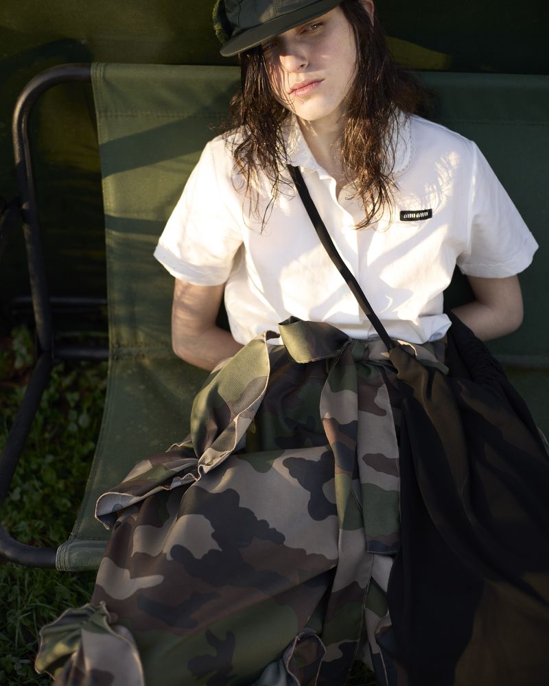 A young adult woman sits on a green chair outside, wearing a white collared shirt, camouflage skirt, and cap in soft, natural lighting.