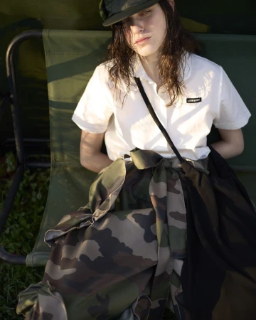 A young adult woman sits on a green chair outside, wearing a white collared shirt, camouflage skirt, and cap in soft, natural lighting.