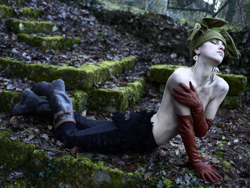 Woman in whimsical bunny hat and gloves poses on mossy stairs in woodland setting, eyes closed in outdoor fashion shot.